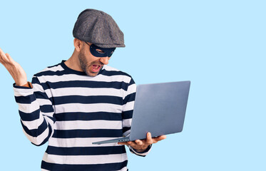 Young handsome man wearing burglar mask using laptop celebrating victory with happy smile and winner expression with raised hands