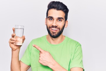 Young handsome man with beard drinking glass of water smiling happy pointing with hand and finger