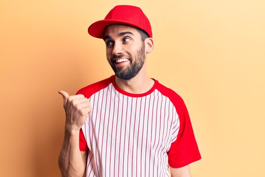 Young handsome man with beard wearing baseball cap and t-shirt pointing thumb up to the side smiling happy with open mouth