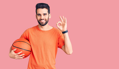 Young handsome man with beard hoilding basketball ball doing ok sign with fingers, smiling friendly gesturing excellent symbol