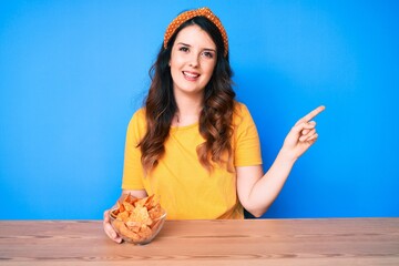 Young beautiful brunette woman sitting on the table eating nachos potato chips smiling happy...