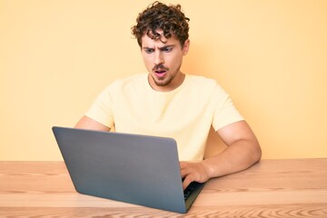 Young caucasian man with curly hair working at the office with laptop scared and amazed with open mouth for surprise, disbelief face