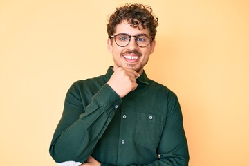 Young caucasian man with curly hair wearing casual clothes and glasses looking confident at the camera with smile with crossed arms and hand raised on chin. thinking positive.