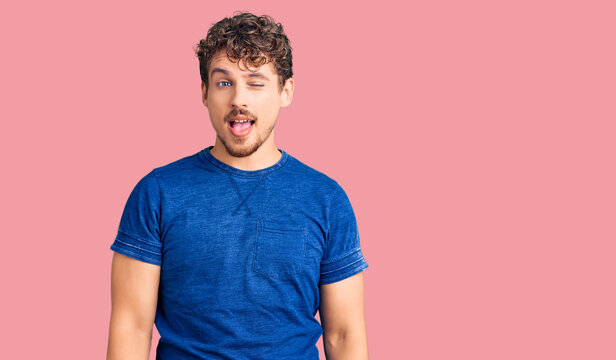Young handsome man with curly hair wearing casual clothes winking looking at the camera with sexy expression, cheerful and happy face.