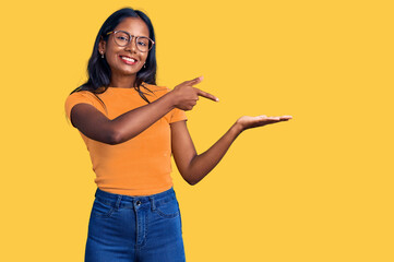 Young indian girl wearing casual clothes and glasses amazed and smiling to the camera while presenting with hand and pointing with finger.