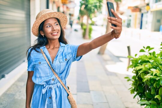 Young beautiful indian woman wearing summer hat smiling happy making selfie by the smartphone at the city.