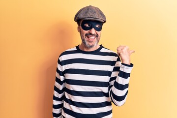 Middle age handsome burglar man wearing cap and mask over isolated yellow background pointing thumb up to the side smiling happy with open mouth