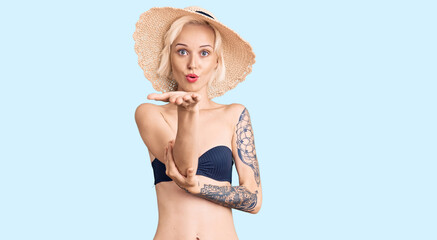 Young blonde woman with tattoo wearing bikini and summer hat looking at the camera blowing a kiss with hand on air being lovely and sexy. love expression.