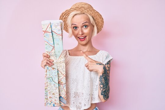 Young blonde woman wearing summer hat holding city map smiling happy pointing with hand and finger