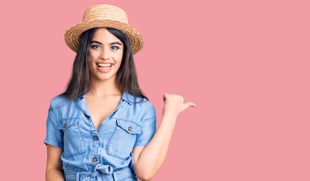 Brunette teenager girl wearing summer hat smiling with happy face looking and pointing to the side with thumb up.