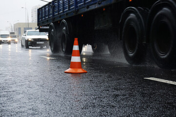 Naklejka na ściany i meble Road cone stands on the road against the background of fast moving cars and a truck in rainy weather.