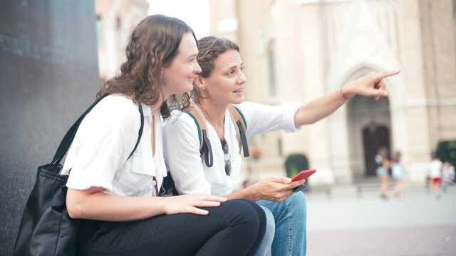 Handheld shot of two young cheerful women sitting on a monument in a city center and looking at a map on a smartphone.