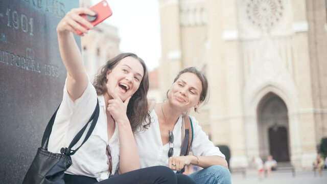 Handheld shot of two young cheerful women sitting on a monument in a city center and taking a selfie with a smartphone.