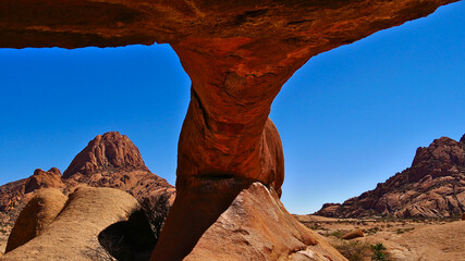 Natural orange colored rock arch with rounded rocks and mountains in background at Spitzkoppe, Kalahari desert, Namibia, Africa with blue sky on sunny day.