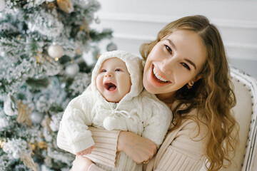 Portrait of a happy mother and a charming child celebrating Christmas. Smiling baby with mom in a festively decorated room with a Christmas tree and decorations.