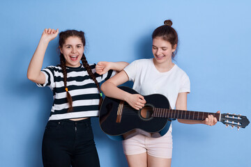 Two friends singing and dancing isolated over blue background, lady with knot playing guitar, winsome girl in striped t shirt and pigtails raising hands up.