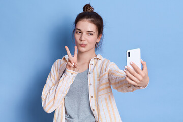 Attractive woman with hair bun in casual outfit showing victory or peace gesture while taking selfie and gesturing v sign, brunette girl against blue background.