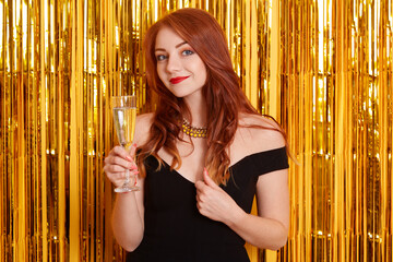 Lady with pleasant appearance looking directly at camera with glass of wine in hands, looking at camera, keeping hand in her hair, wearing black dress.