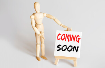 Wooden man shows with a hand to white easel with text COMING SOON