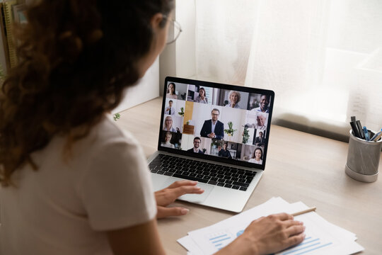 Rear view of female employee at home talk on team video call on laptop, engaged in group online meeting or briefing. Woman worker have webcam digital virtual conference with diverse colleagues on web.