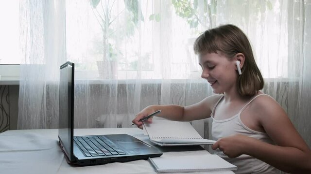 little girl wears headphones, studies at home, writes notes in notebook, shows copybook to somebody. schoolgirl listens to tutor, doing homework. online education, video call tutoring