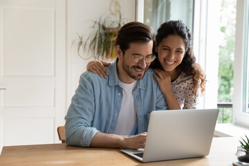 Happy Caucasian young couple look at laptop screen browse surf wireless internet on gadget. Smiling millennial man and woman shopping paying on internet on computer at home. Technology concept.