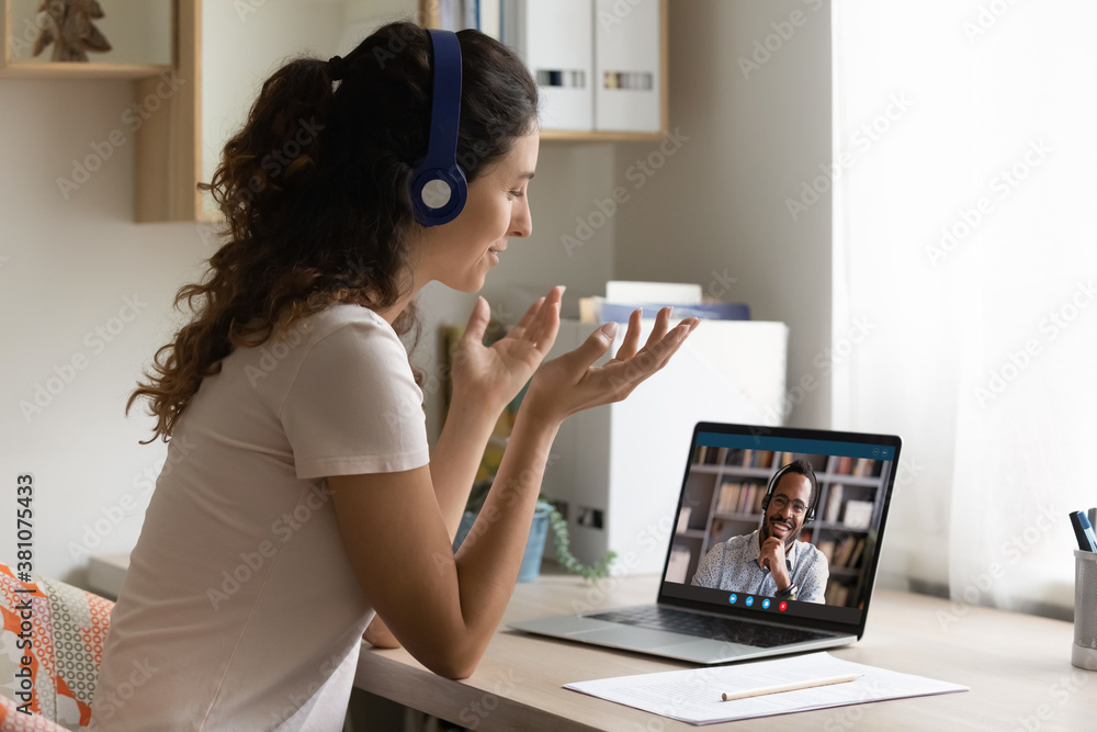 Sticker Happy Caucasian young woman look at laptop screen talk chat on video call with African American man husband. Smiling female in headphones have webcam digital virtual communication with friend.