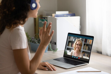 Close up side view of happy woman in headphones look at laptop screen wave greet talk on video call with mature mother. Smiling young female in earphones have pleasant webcam conference on computer.