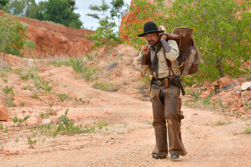A cowboy man walks saddles along a small dirt road in a mine.