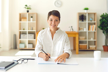 Smiling young woman taking notes while listening to an online lecture in a bright room.