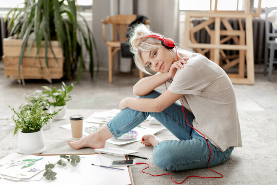 Creative beautiful young woman artist wearing headphones connected to smartphone, casual clothes sitting sideways and looking at camera on floor in modern art studio. Break and relax time