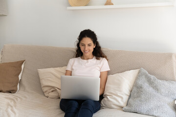 Smiling young Caucasian woman sit relax on comfortable couch in living room use laptop at home. Happy millennial female renter tenant rest on sofa, browse wireless internet on computer online.