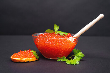 National Russian holiday delicacy - red salmon caviar in glass bowl with wooden spoon and on piece of bread decorated with fresh parsley, black background, front view.