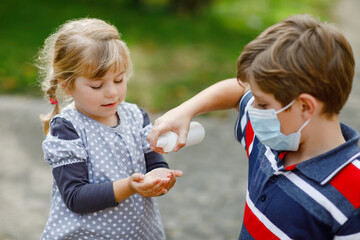 School kid boy helping little toddler sister cleaning hands with sanitizer spray. Brother and cute little girl learn hygiene rules. Family during coronavirus and flu lockdown. Covid virus protection.