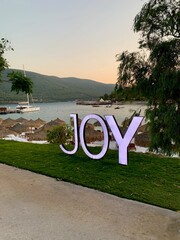 Fototapeta premium Large letters: JOY, Panoramic view of the emerald Aegean coast with white sand, bamboo palms and sun loungers. Luxury leisure relaxation concept. White beach with a mountain in the background
