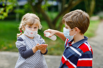 School kid boy helping little toddler sister cleaning hands with sanitizer spray. Brother and cute little girl in medical mask learn hygiene rules. Family during coronavirus pandemic quarantine.