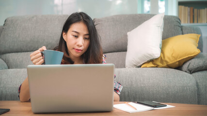 Naklejka premium Young business freelance Asian woman working on laptop checking social media and drinking coffee while lying on the sofa when relax in living room at home. Lifestyle women at house concept.