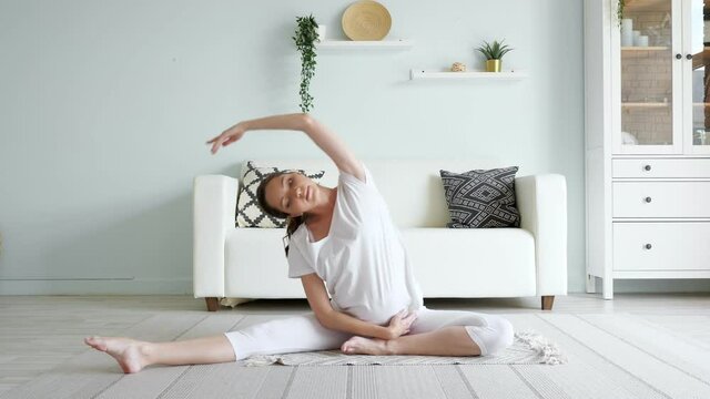 Young pregnant lady in white clothes does yoga janu sirsasana sitting on floor with rug against designer sofa in spacious room at home