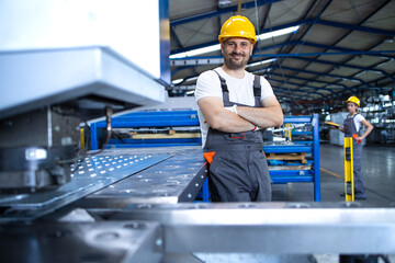 Portrait of factory worker in protective uniform and hardhat standing by industrial machine at...