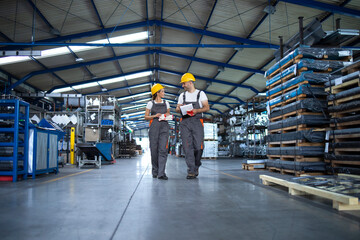 Factory workers in work wear and yellow helmets walking through industrial production hall and...