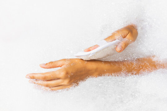 Woman washing hands in bath tub with foam bubbles and use natural jute sponge. Hands close-up. Spa treatment, body care.