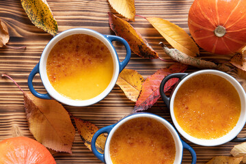 orange Creme Brulee aus Kürbis vom Hokkaido mit  karamellisierter Zuckerkruste in Förmchen auf einem braunen Holztisch mit Laubblättern im Herbst - obrazy, fototapety, plakaty