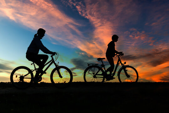 Boy , kid 10 years old, and girl riding bikes in countryside in amazing colorful cloudy night sky background, silhouette of riding persons at sunset in nature 