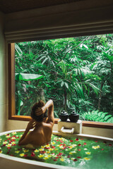 Beautiful young woman enjoying in spa, view from behind. Luxury stone bath tub with jungle view in window. Tropical flowers in water. Beauty treatment concept.