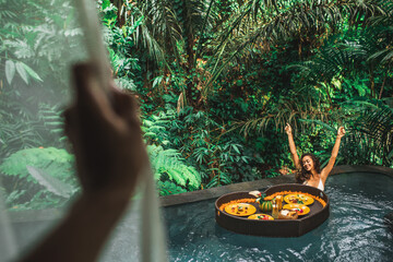 Girl relaxing and eating floating breakfast in jungle pool on luxury villa in Bali. View through...