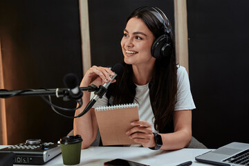 Portait of attractive young female radio host smiling aside while speaking in microphone,...