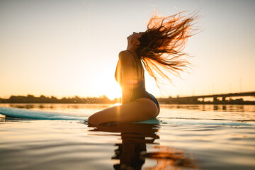 side view of woman with long flowing hair sitting on surfboard against backdrop of rising sun