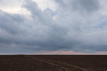Fototapeta premium Autumn cultivated land rural bare field with dirt road going to horizon with dramatic sunrise colored gorgeous clouds. Plowed field with grey epic sunset layered cloudscape