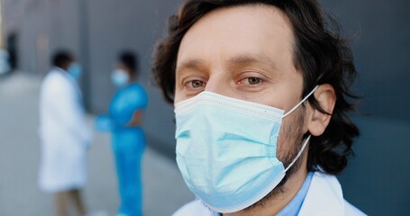 Portrait of Caucasian handsome man doctor in medical mask looking at camera. Close up of male physician in respiratory protection. African American medics colleagues on background.