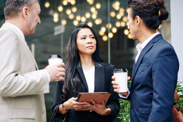 Serious young Asian business lady with tablet computer looking at her talking colleagues when they are standing outdoors
